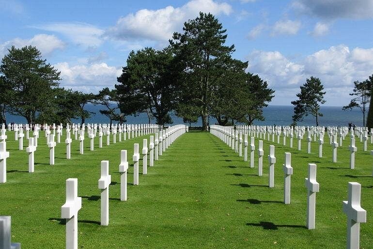 Rows of white marble crosses on a lawn under pine trees, the sea beyond